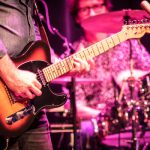 Musician playing an electric guitar on stage with a drummer visible in the background.
