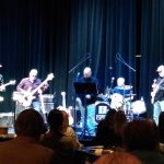Band performing live on a dark stage with musicians and audience members watching.