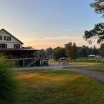 Rustic lodge by a green field at sunset with trees lining the horizon.