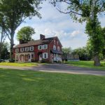Historic brick house surrounded by mature trees on a sunny, green lawn.