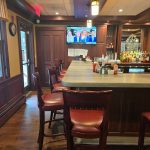 Bar area with wooden counter, stools, and a television displaying people.