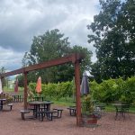 Wooden pergola over a gravel area with tables and chairs, set against a backdrop of lush green vineyards.