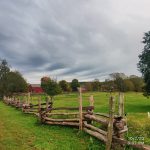 Rustic wooden fence borders a green field under an overcast, cloudy sky with distant trees and barns.