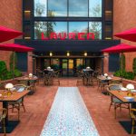 Outdoor patio dining area in front of a modern brick building with red umbrellas.