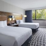 Modern hotel room with two white beds, wood accents, and large window view.
