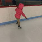 Child in pink coat skates on an indoor ice rink.