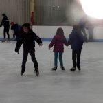 Children ice skating indoors in a rink, some wearing dark coats.