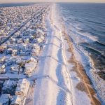 Snow-covered coastal town meets a wide, wintry beach with ocean waves.