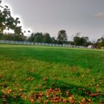 Green field with white fence under a cloudy sky, trees in background.