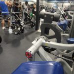 Weightlifting gym interior with exercise equipment and people working out in the background.