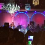 Live band performance on stage with colorful lighting and a crowd watching.