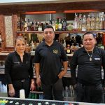 Three men stand in front of a well-stocked bar area.