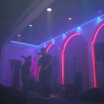 Live band performing on a dimly lit stage with colorful spotlights and dramatic lighting.