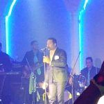 Male singer performs on stage under bright blue arch lighting with a band.
