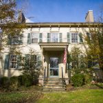 Historic two-story colonial house with green shutters, set on a grassy lawn under blue sky.