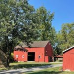 Red barn and stone outbuilding under green trees on a sunny day.
