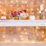 Wedding display with cakes, flowers, and string lights on a rustic wooden table.