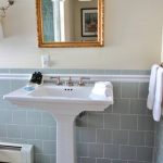 White pedestal sink in a bathroom with grey subway tile and gold mirror.