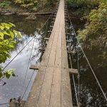 Suspension bridge crosses a dark river surrounded by lush green trees.