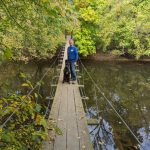 Man and dog walk across a wooden suspension bridge over a calm, wooded pond.