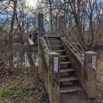 Concrete footbridge crosses a muddy stream through bare winter trees.