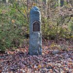 Stone monument in a wooded area with fallen autumn leaves on the ground.