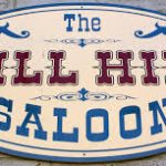 Vintage oval sign reading "The Mill Hill Saloon" in rustic lettering.