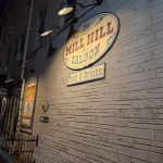 "The Mill Hill Saloon" sign on a textured brick building at night.
