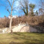 Stone staircase leading up a wooded hill toward a white house under a blue sky.