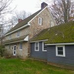 Stone and clapboard farmhouse with a mossy roof, set on a grassy lawn under bare trees.