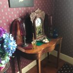 Antique wooden console table with a clock, flowers, and decorative items against patterned wallpaper.