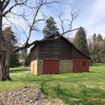 Rustic wooden barn with stone base sits in a grassy field under a bright sky.