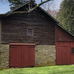 Rustic, dark wood barn with stone foundation and red doors set against a green lawn.