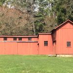 Red barn complex with multiple sections set against a backdrop of trees and green lawn.