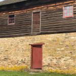 Rustic wooden and stone barn with red trim windows and a central door.