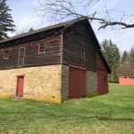 Old wooden barn with stone base in a grassy field under a partly cloudy sky.