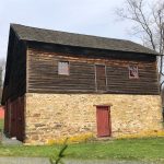 Rustic, dark wood barn with a stone foundation stands in a grassy field under a light sky.