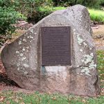 Stone monument with a bronze plaque dedicated to John Hart, set in a grassy outdoor area.