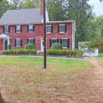 Red brick farmhouse with dark shutters, surrounded by trees and a grassy yard.