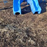 Young child slides down blue playground equipment on a dirt surface outdoors.