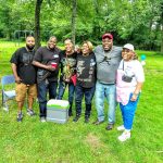 Group of diverse people posing outdoors in a grassy, wooded area.