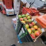 Boxes of produce, including apples and oranges, stacked outdoors near shipping containers.