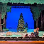 Stage set with trees and a blue backdrop, featuring a Christmas tree and seated audience members.