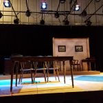 Stage set with a wooden table and chairs under dramatic theatrical lighting.