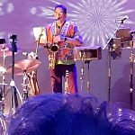 Musician playing trumpet on stage with drums and bright lighting backdrop.