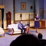 Actors on a stage set in a period room, one woman standing and another seated.