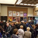 Crowd gathers inside a theater lobby with posters and box office visible.