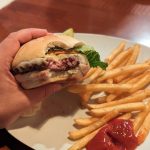 Hand holding a burger next to a plate of french fries and ketchup.