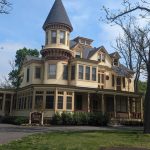 Victorian-style house with a turret under a bright blue sky, surrounded by trees.