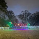 Holiday light display spelling "SEASONS GREETINGS" on a grassy field at dusk.
